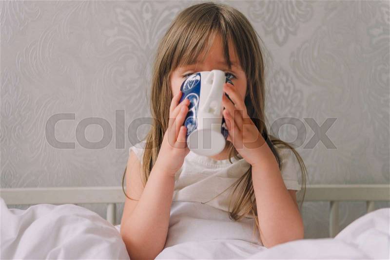 Little girl in the bedroom on the bed. Little girl wearing a pajamas sitting on a bed and drinking water from a cup. Attractive girl in a pajamas holding a cup sitting in white bedding, stock photo