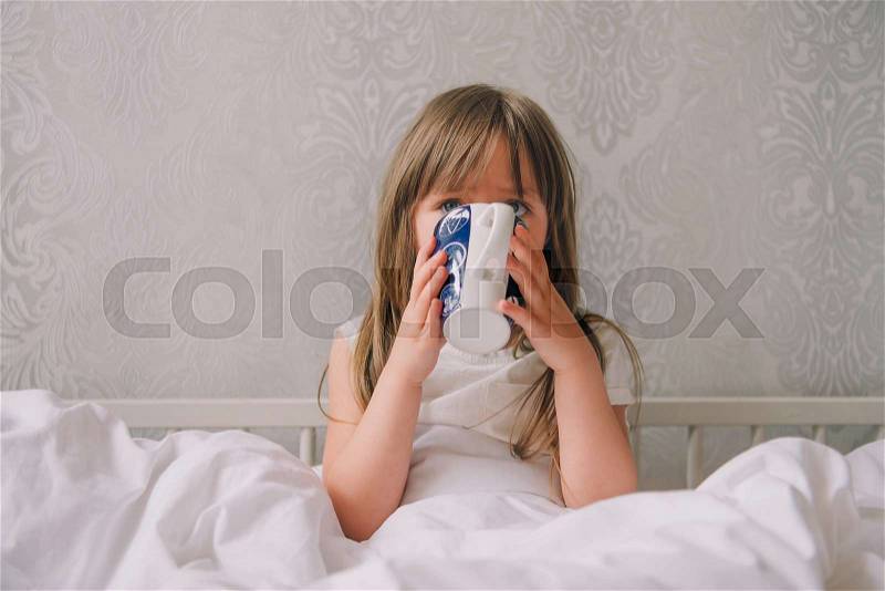 Little girl in the bedroom on the bed. Little girl wearing a pajamas sitting on a bed and drinking water from a cup. Attractive girl in a pajamas holding a cup sitting in white bedding, stock photo