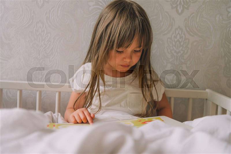 Little girl reads the book. small girl sits in bed and sees a book. little girl is considering a book. child reads and watches pictures in the book, stock photo