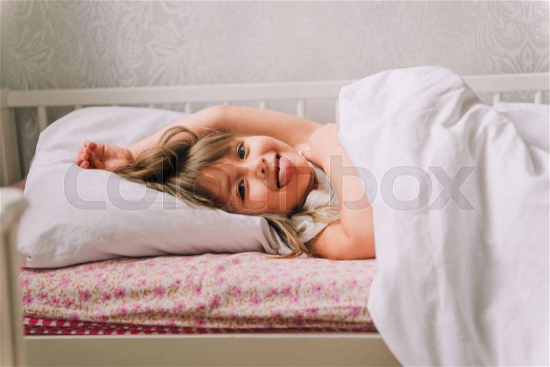 Little girl lying in bed. Little girl dressed in pajamas, lying in bed and smiling. next to the bedside table is an alarm clock and cup, stock photo