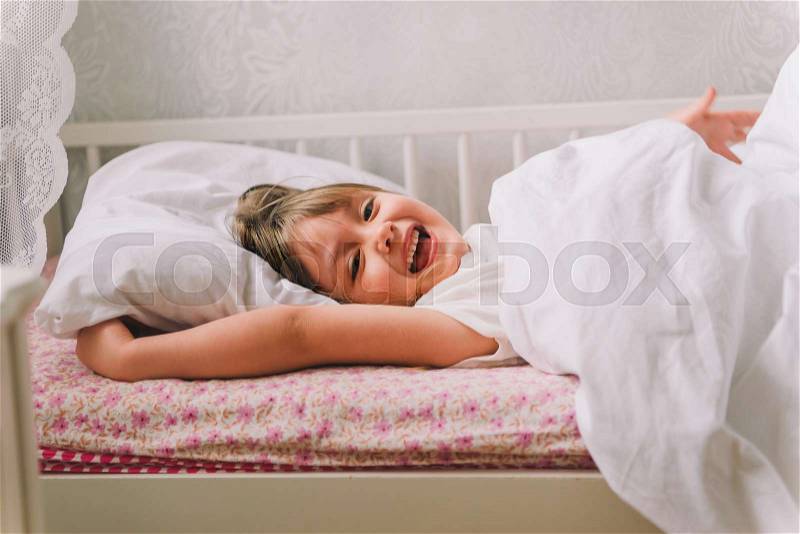 Little girl lying in bed. Little girl dressed in pajamas, lying in bed and smiling. next to the bedside table is an alarm clock and cup, stock photo