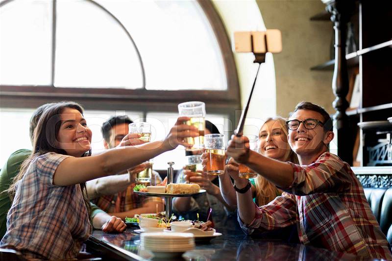 People, leisure, friendship and technology concept - happy friends taking picture by smartphone selfie stick, drinking beer and eating snacks at bar or pub, stock photo