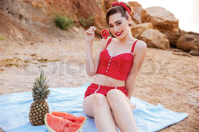 Smiling pin-up girl having great time at the beach holding heart shaped candy, stock photo