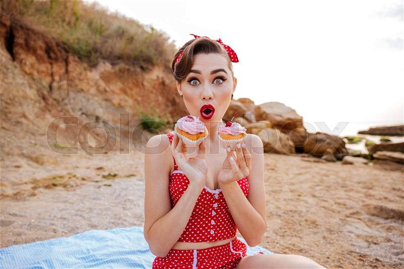 Surprised young pin up girl showing two cream cakes sitting outdoors, stock photo