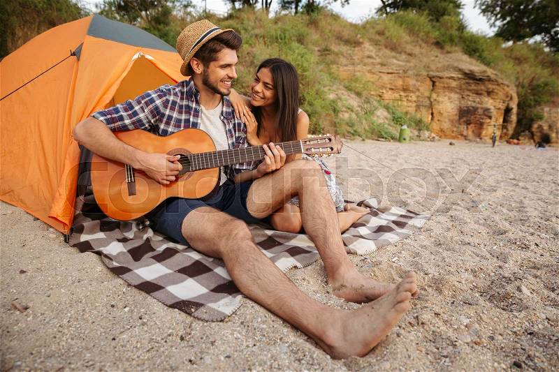 Young beautiful happy couple playing guitar while sitting at the beach, stock photo