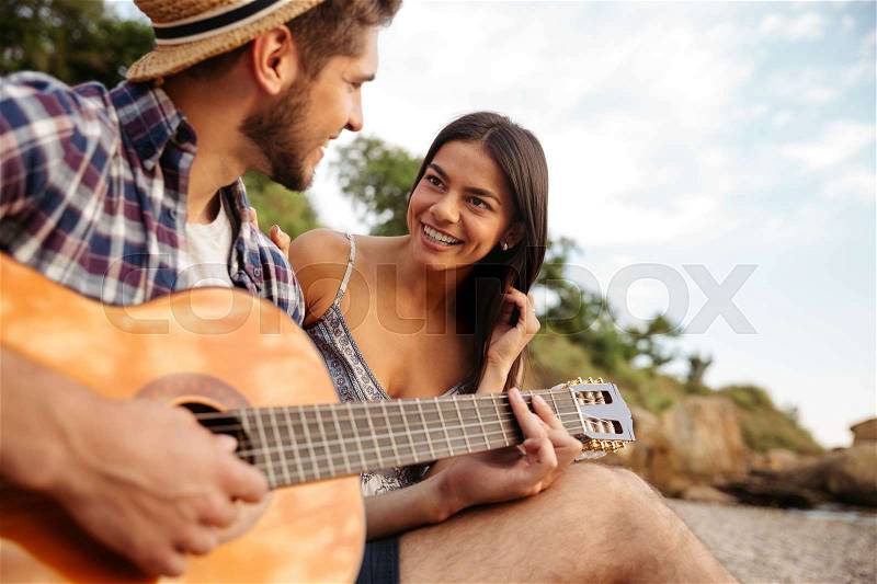 Young beautiful happy couple playing guitar while sitting at the beach, stock photo
