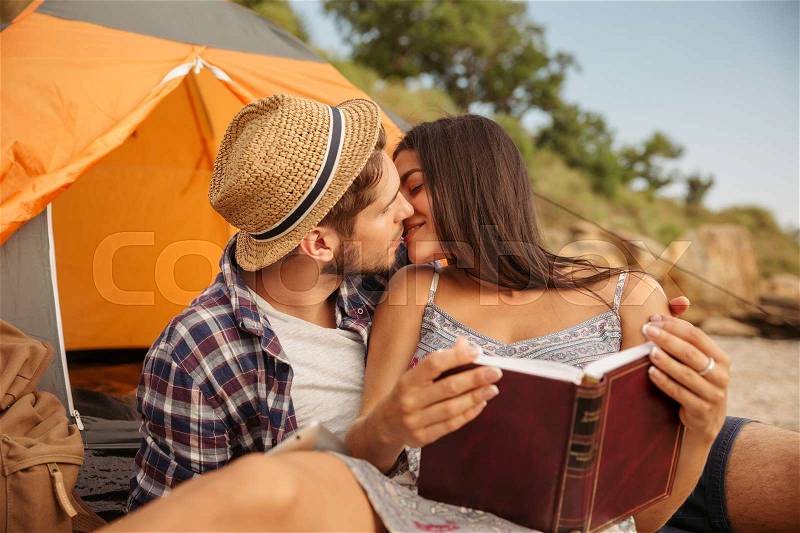 Portrait of a happy couple sitting at the tent and kissing while using tablet computer and reading book, stock photo
