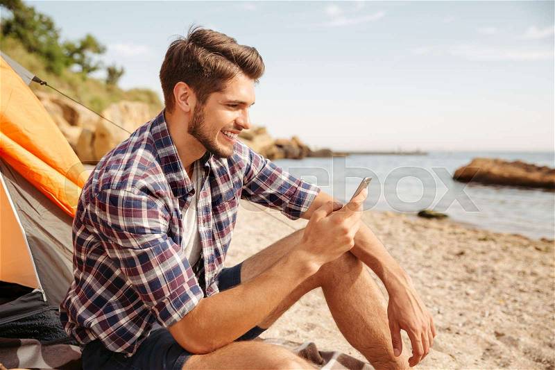 Smiling young man tourist using smartphone sitting in touristic tent at the beach, stock photo