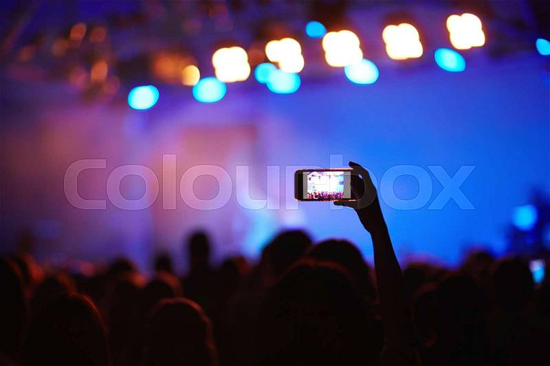 Back lit image of crowd at music concert before stage, focus on one fan holding cellphone high above heads to take picture of memorable moment, stock photo