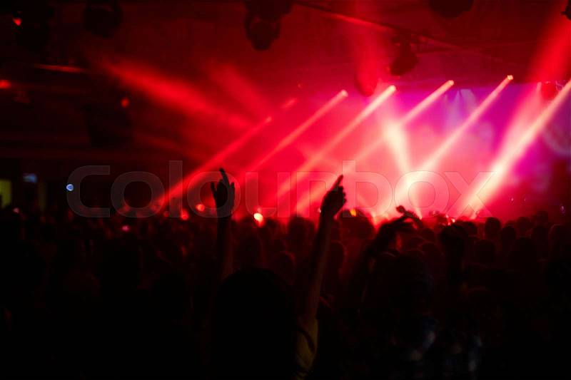 Large crowd of cheering excited fans dancing and jumping in front of big stage in rays of red light, stock photo