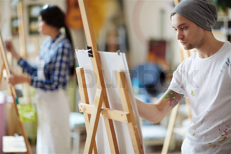 Male artist dressed in beanie hat and white shirt covered in colorful paint sketching on easel in brightly lit studio with blurred female artist in apron in background, stock photo