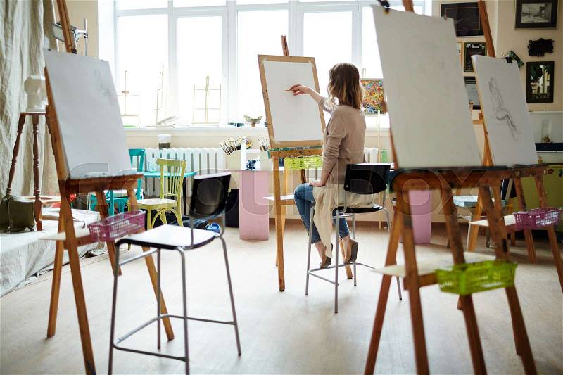 Rear view of young woman sitting near window sketching on easel with pencil in brightly lit studio, stock photo