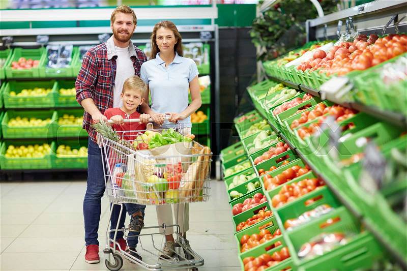 Happy consumers with push-cart looking at camera in hypermarket, stock photo