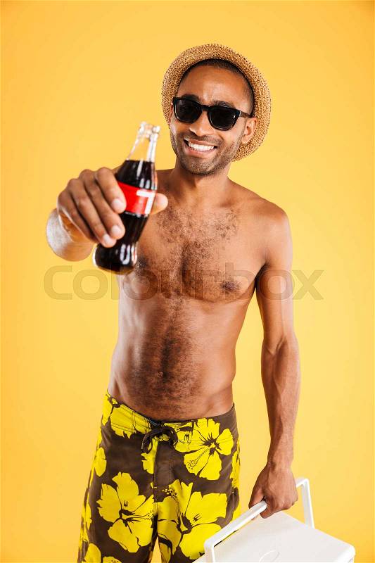 Cheerful young man in hat and sunglasses holding coke bottle and cooler bag over orange background, stock photo
