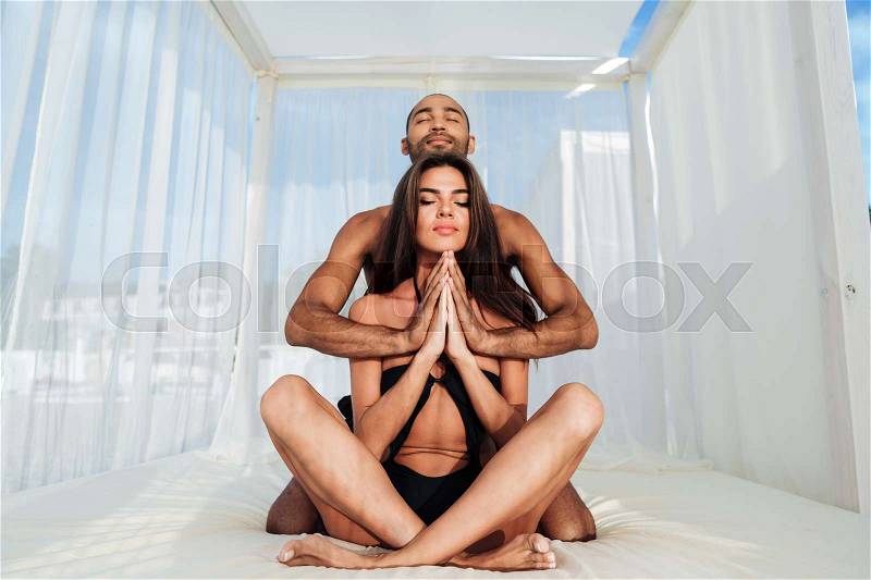 Beautiful multiracial couple mediating together and sitting in lotus position on the beach bed, stock photo