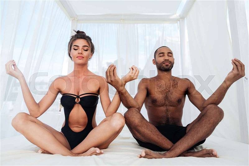 Beautiful multiracial couple mediating together and sitting in lotus position on the beach bed, stock photo