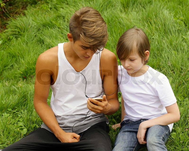 Older and younger brother sitting on the grass and looking to the smartphone, stock photo