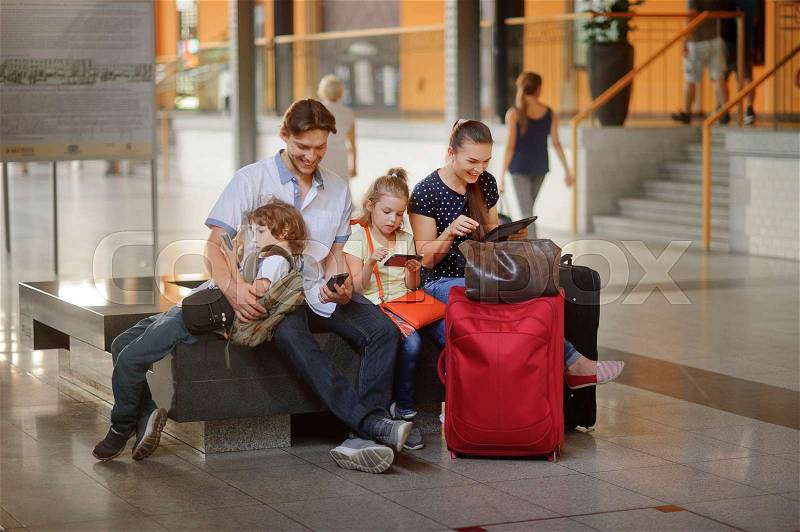 Young parents with two children at the railway station. Family in a large waiting room. All have settled down on a bench. Everyone looks in your gadget. Nearby are the suitcases and travel bags, stock photo