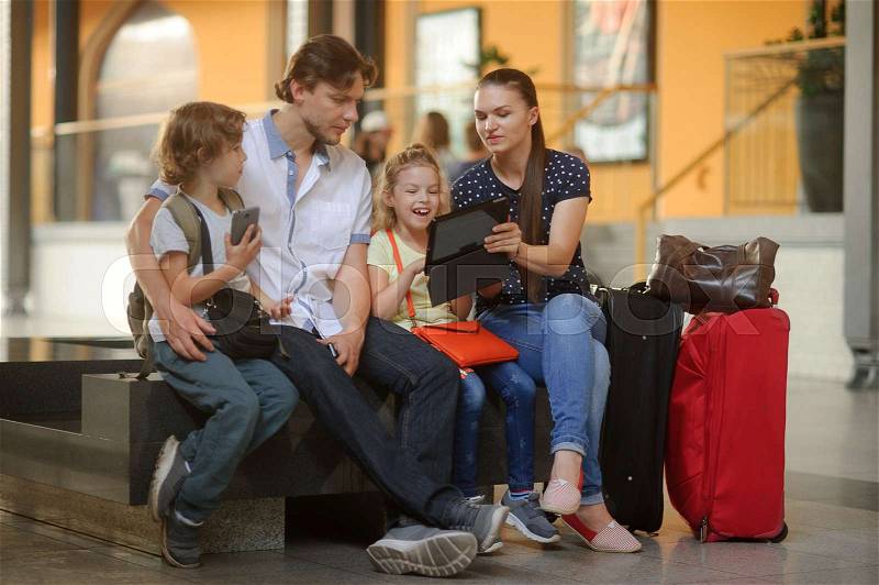 Parents with two children at the railway station. Family in large waiting room. All have settled down on bench. Mom shows something interesting on the screen of the tablet. Nearby are the travel bags, stock photo