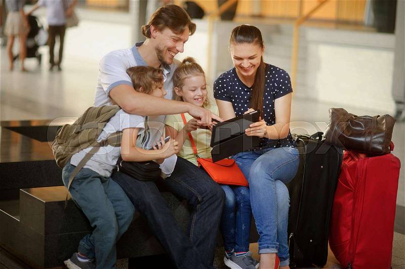 Parents with two children at the railway station. Family in large waiting room. All have settled down on bench. Mom shows something interesting on the screen of the tablet. Nearby are the travel bags, stock photo