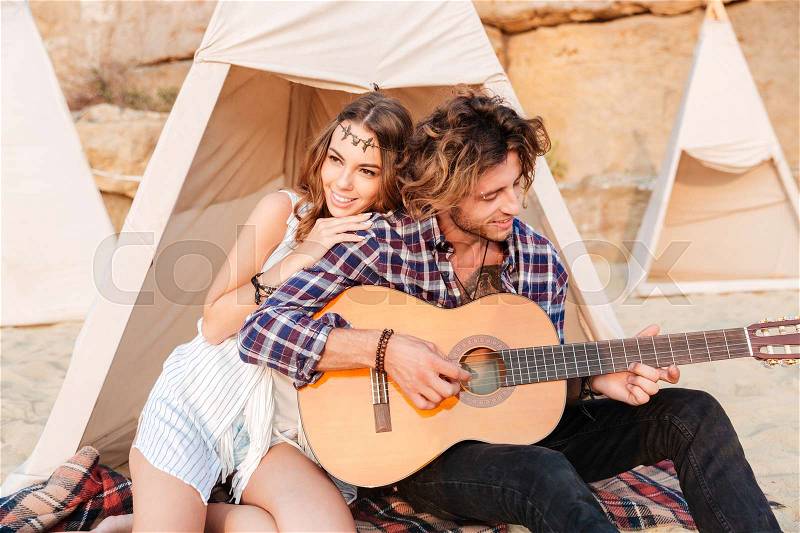 Curly young man playing guitar for his girlfriend sitting at the camping tent, stock photo