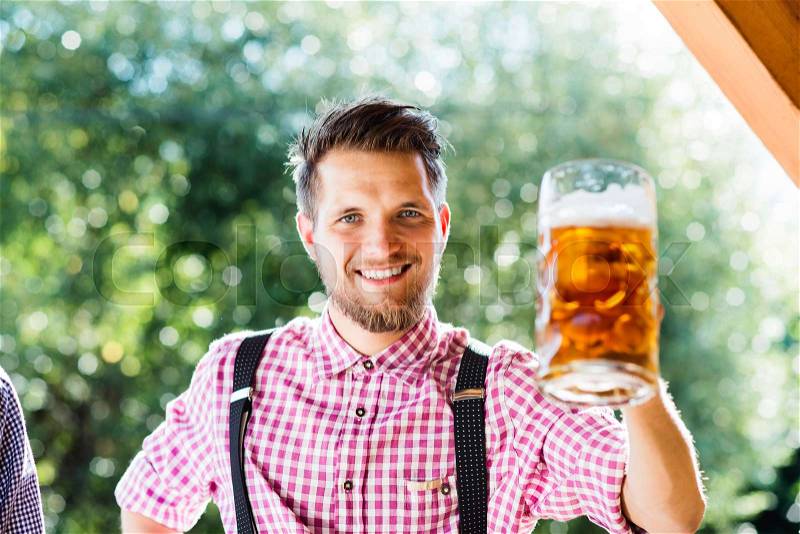 Handsome hipster young man in traditional bavarian clothes holding a mug of beer. Oktoberfest. Sunny summer garden, stock photo