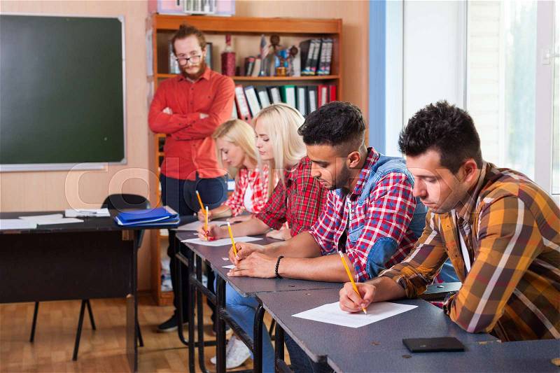 Student Group Write Test, Professor Observing, Young Diverse People Sit Desk University Classroom Examination High Shool Education, stock photo