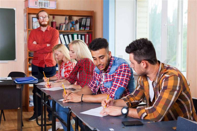 Student Group Write Test, Professor Observing, Young Diverse People Sit Desk University Classroom Examination High Shool Education, stock photo