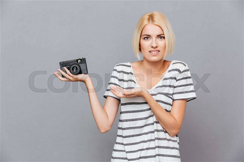 Confused blonde young woman holding old vintage photo camera over gray background, stock photo