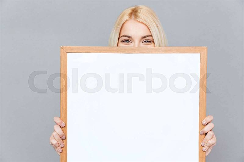 Cute blonde young woman hiding her face behind blank white board over gray background, stock photo