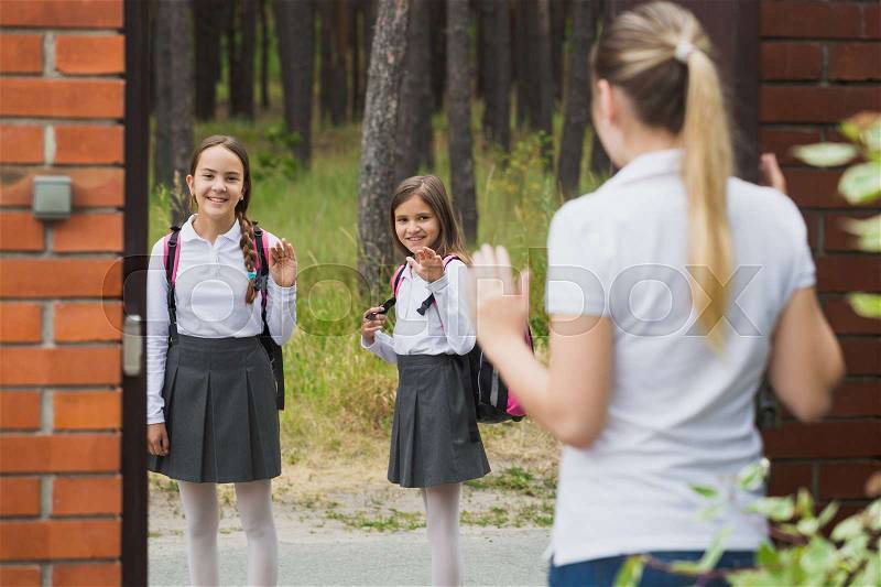 Young mother standing in house yard and waving to her daughters walking to school, stock photo
