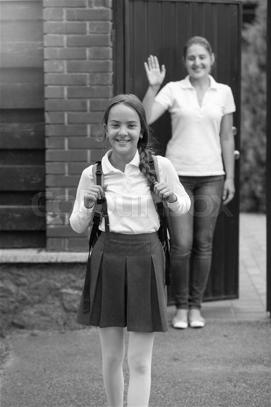 Black and white image of beautiful smiling girl in schoolgirl standing in front of mother waving to her, stock photo