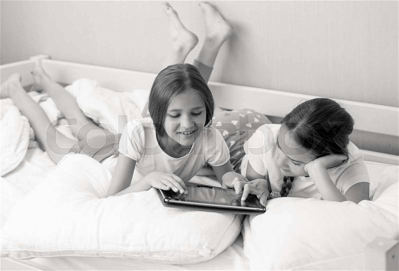 Black and white portrait of two teenage sisters using tablet on bed at morning, stock photo