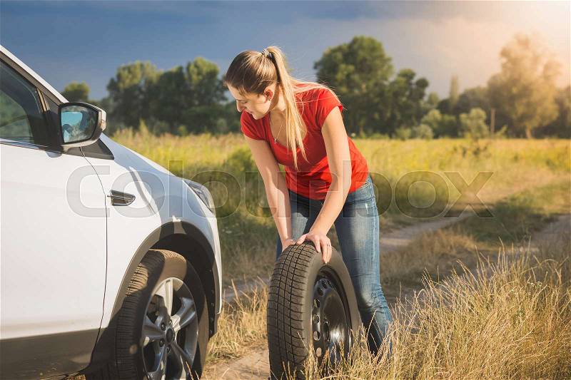 Beautiful young woman rolling spare tire to change the flat one, stock photo