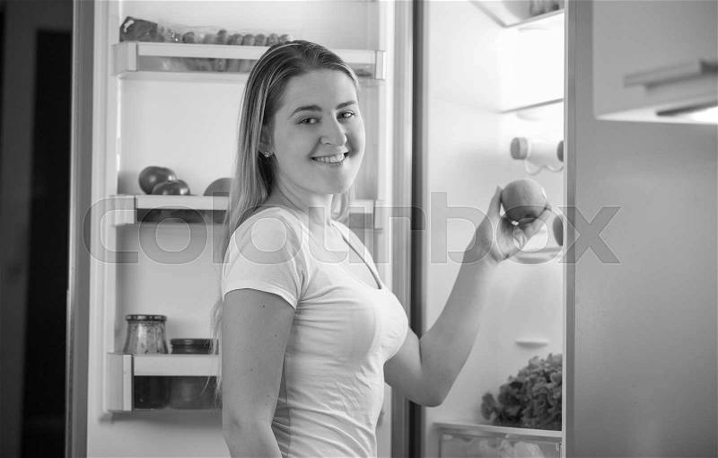Black and white portrait of young hungry woman taking apple from refrigerator at night, stock photo