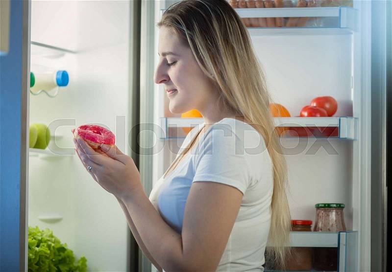 Portrait of young woman taking donut from refrigerator at night, stock photo