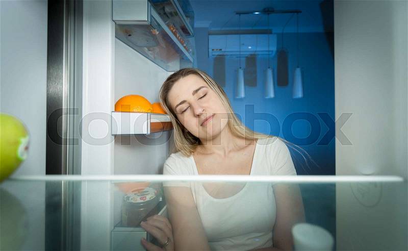 Portrait of tired woman in pajamas looking inside of refrigerator at late night, stock photo
