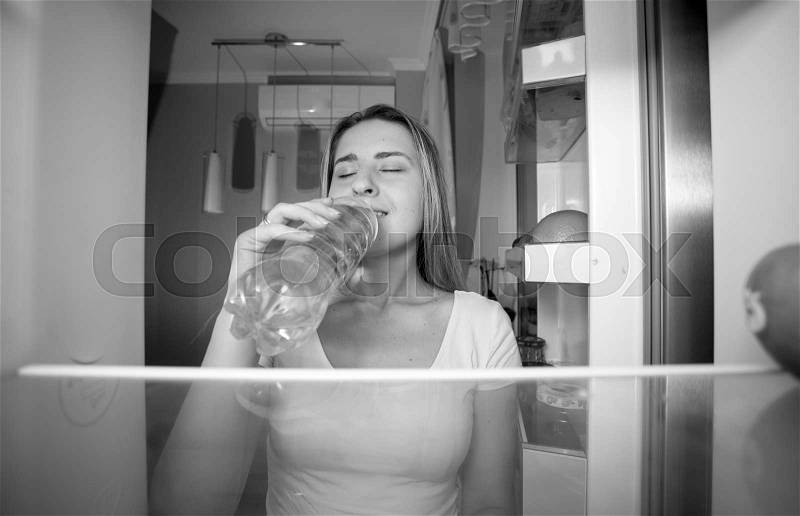 Black and white portrait of beautiful smiling woman taking water from fridge and drinking it. View from inside of open refrigerator, stock photo