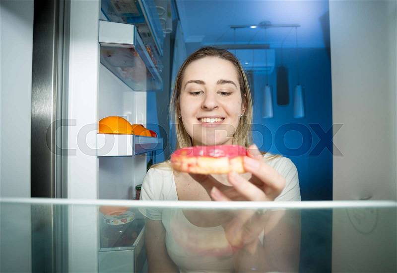 Portrait of young woman opening refrigerator and taking big donut at night, stock photo