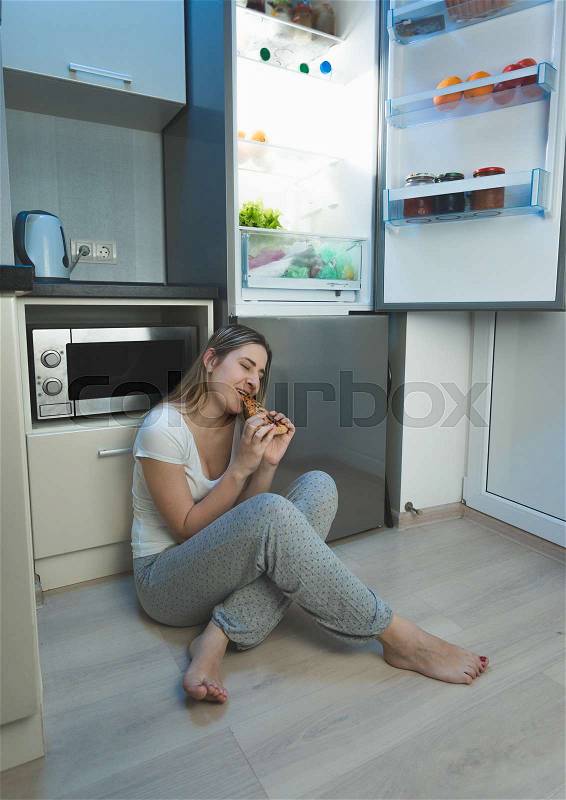 Young sleepless woman sitting on kitchen floor next to open refrigerator and eating pizza, stock photo