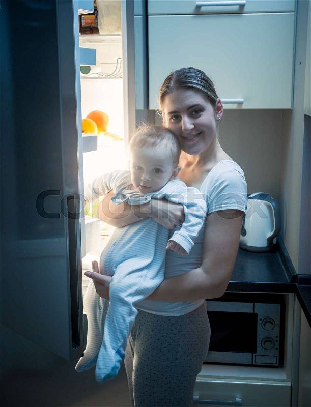 Young smiling woman standing with her baby son on kitchen at night, stock photo
