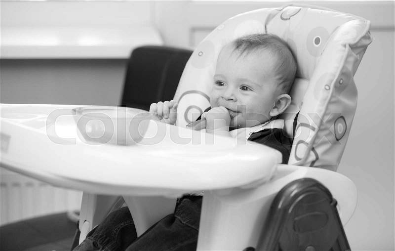 Black and white portrait of 9 months old baby sitting in highchair and playing with food, stock photo