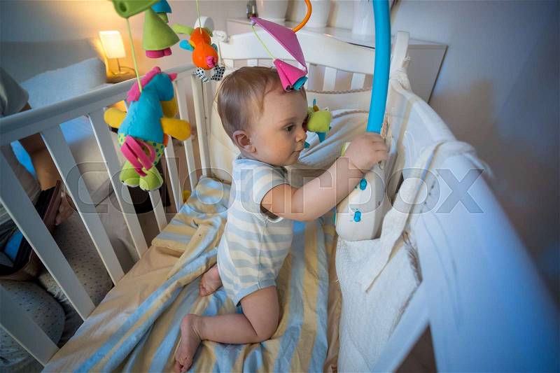 Portrait of adorable baby boy standing in crib and playing with toy carousel, stock photo