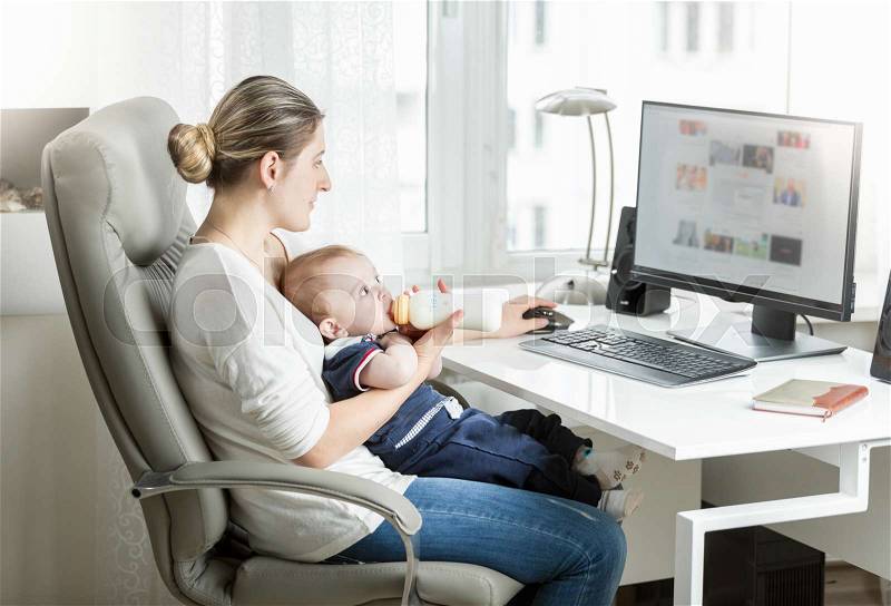 Beautiful smiling mother working at home office and feeding her baby from bottle, stock photo
