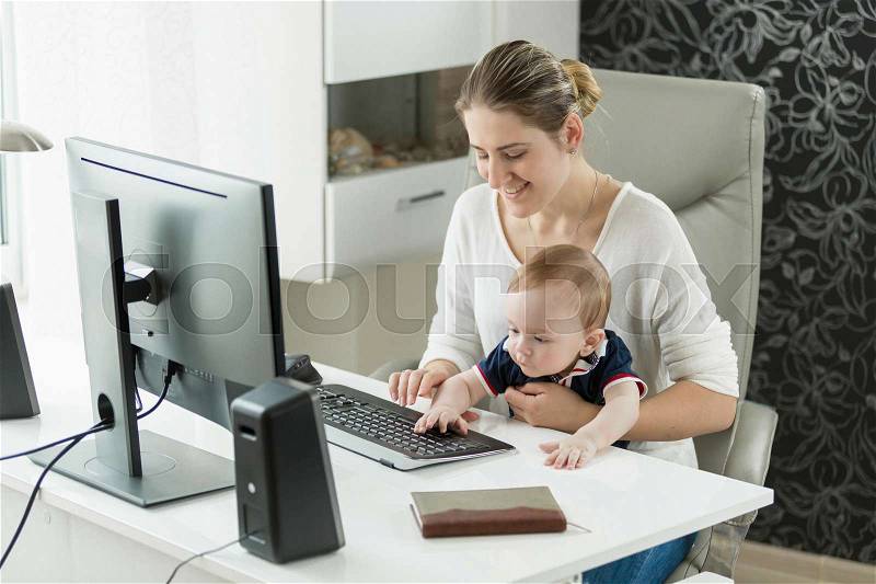 Portrait of smiling businesswoman working at computer with baby boy sitting on her lap, stock photo