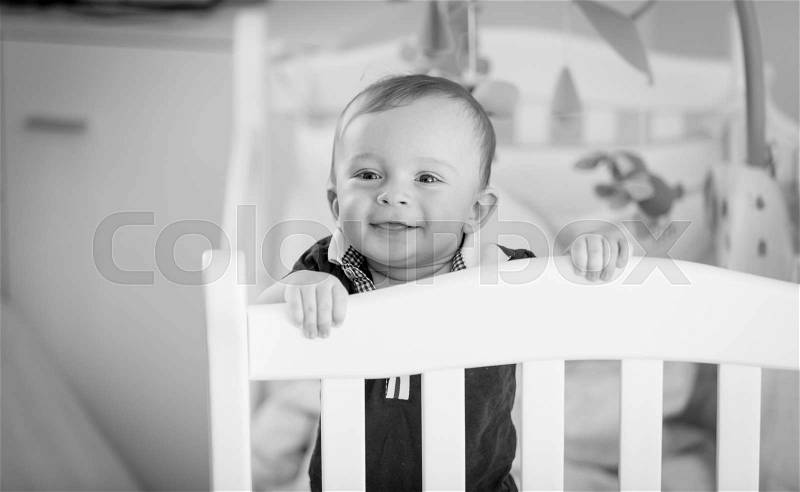 Black and white portrait of cute baby boy standing at his crib, stock photo