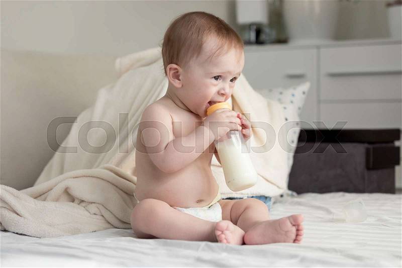 Beautiful 9 months old baby boy sitting on bed and drinking milk from bottle, stock photo