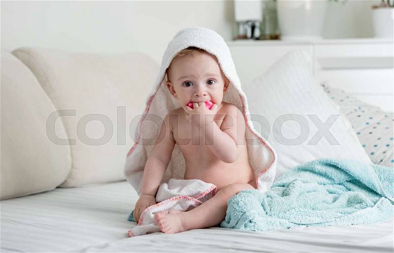 Adorable 9 months old baby boy sitting on bed under towel, stock photo