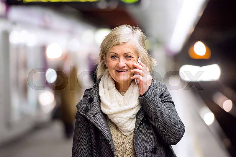 Beautiful senior woman standing at the underground platform, waiting, talking on the phone, stock photo