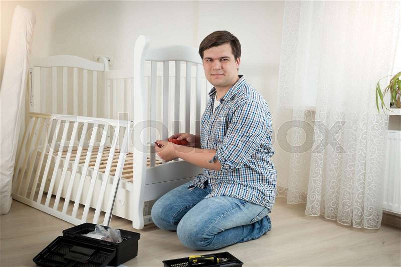 Happy young man assembling white wooden crib in nursery, stock photo